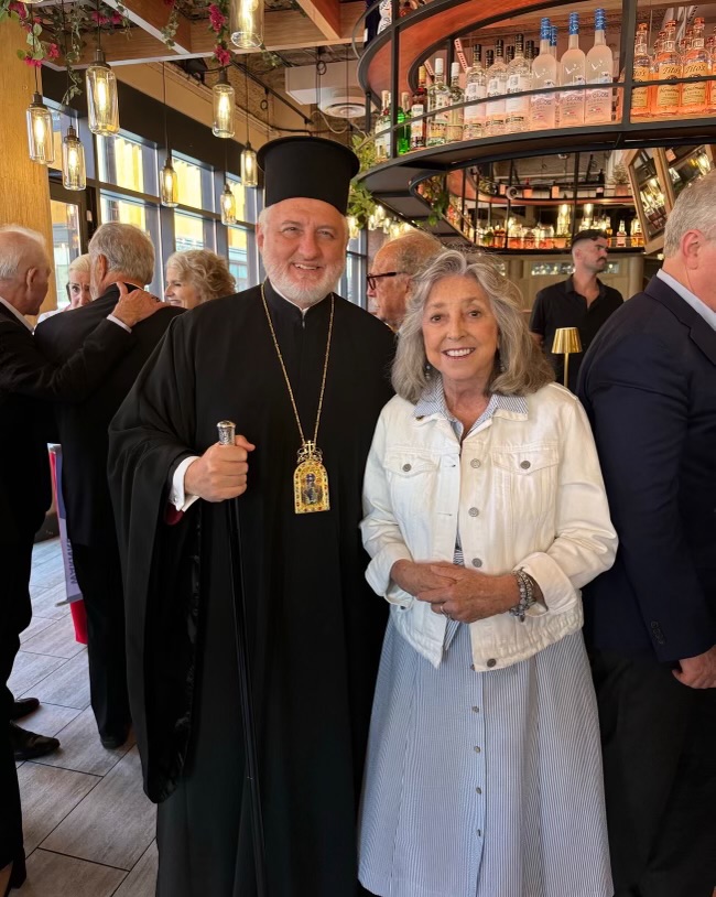 Rep. Titus in a white jacket with a long blue skirt standing next to the Archbishop in traditional attire